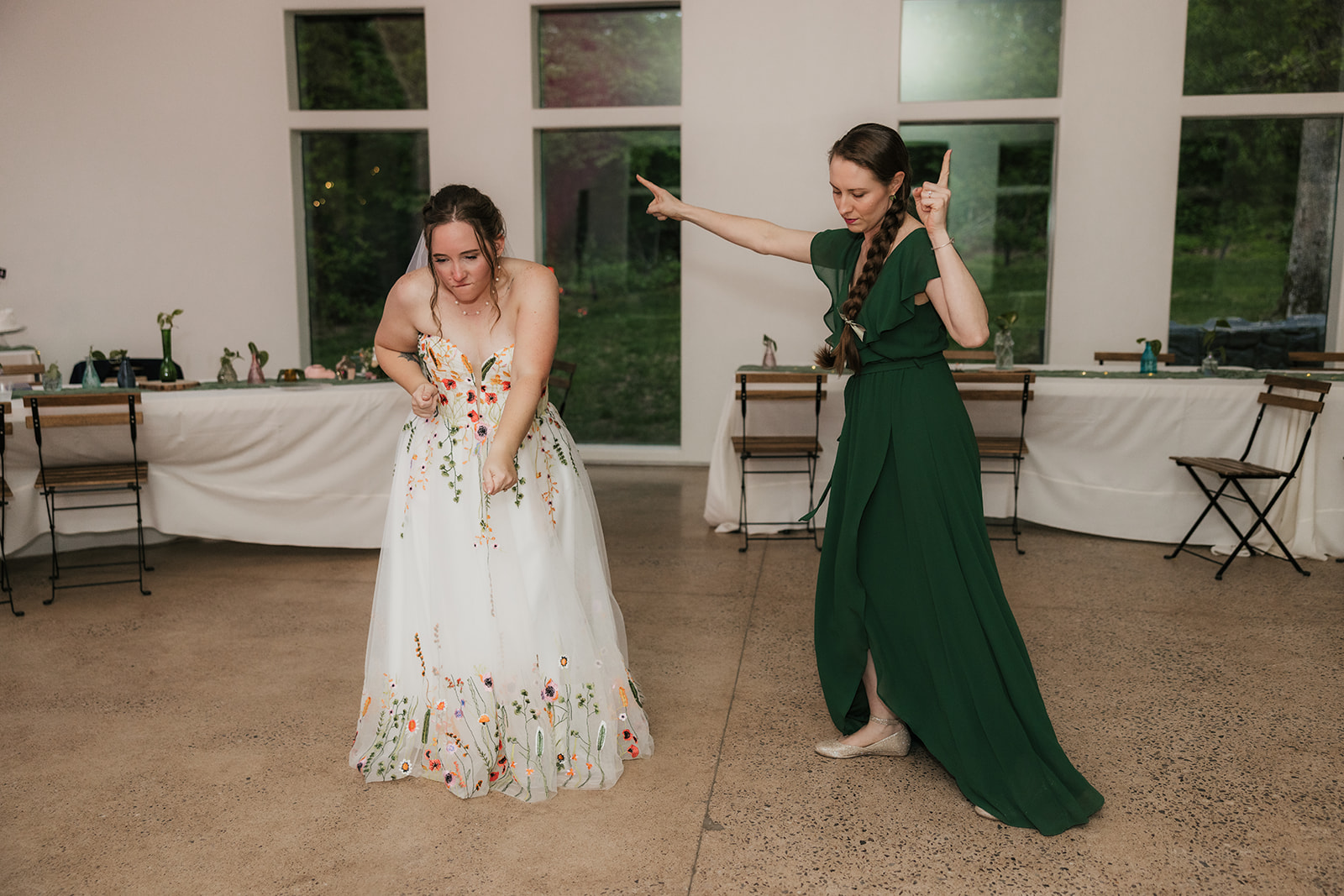 Two women dance indoors; one wears a white floral dress, the other a green dress. They appear to be enjoying themselves in a room with large windows and simple decor for their wedding at Merrick Hollow 