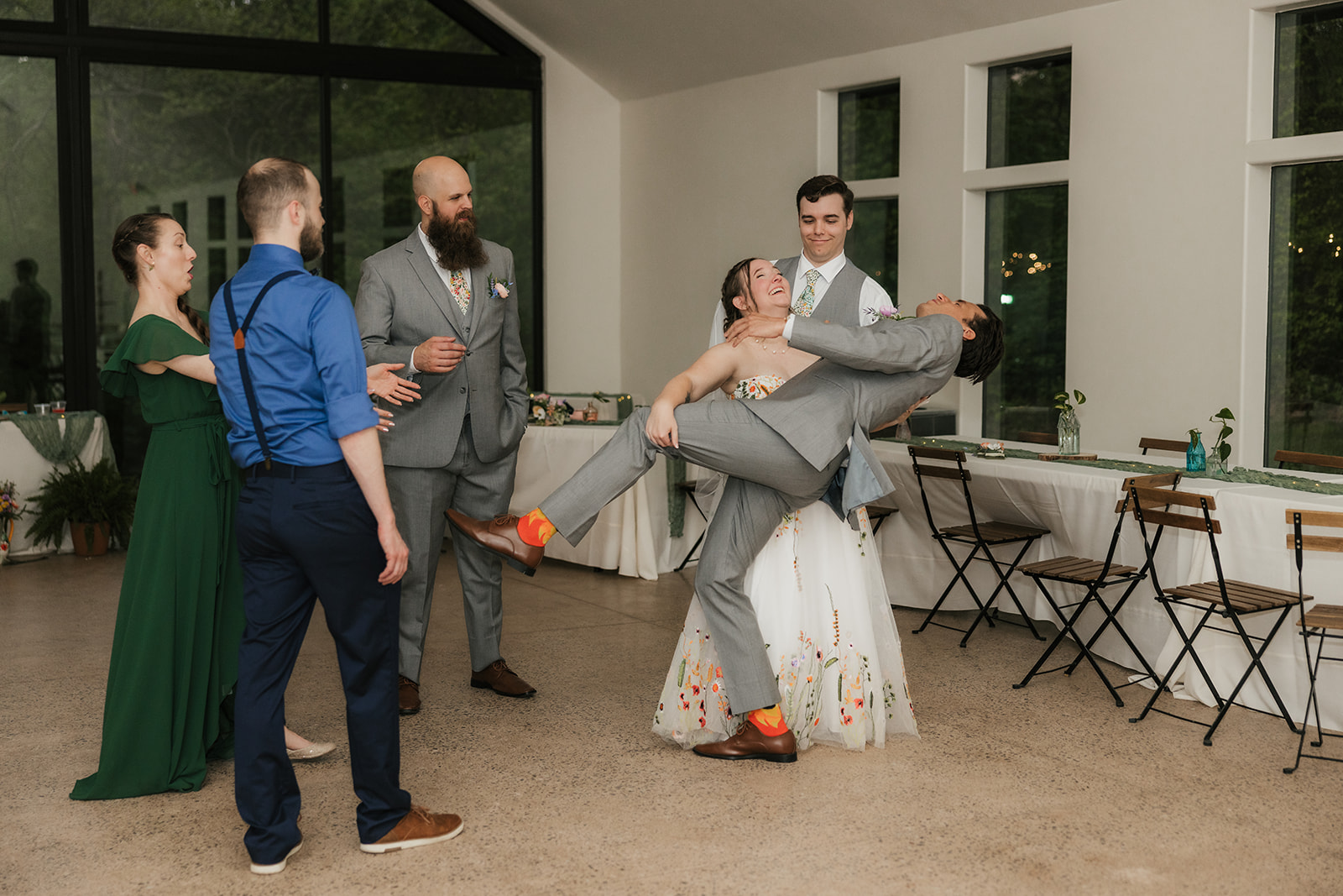 Two women dance indoors; one wears a white floral dress, the other a green dress. They appear to be enjoying themselves in a room with large windows and simple decor for their wedding at Merrick Hollow 