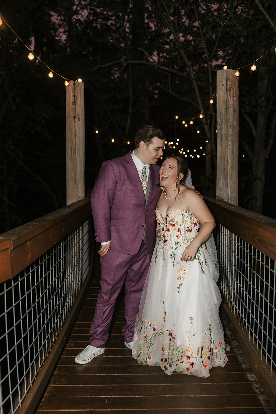 A couple stands on a wooden bridge under string lights at dusk; the person on the left wears a purple suit, and the person on the right wears a white floral dress for their wedding at Merrick Hollow 