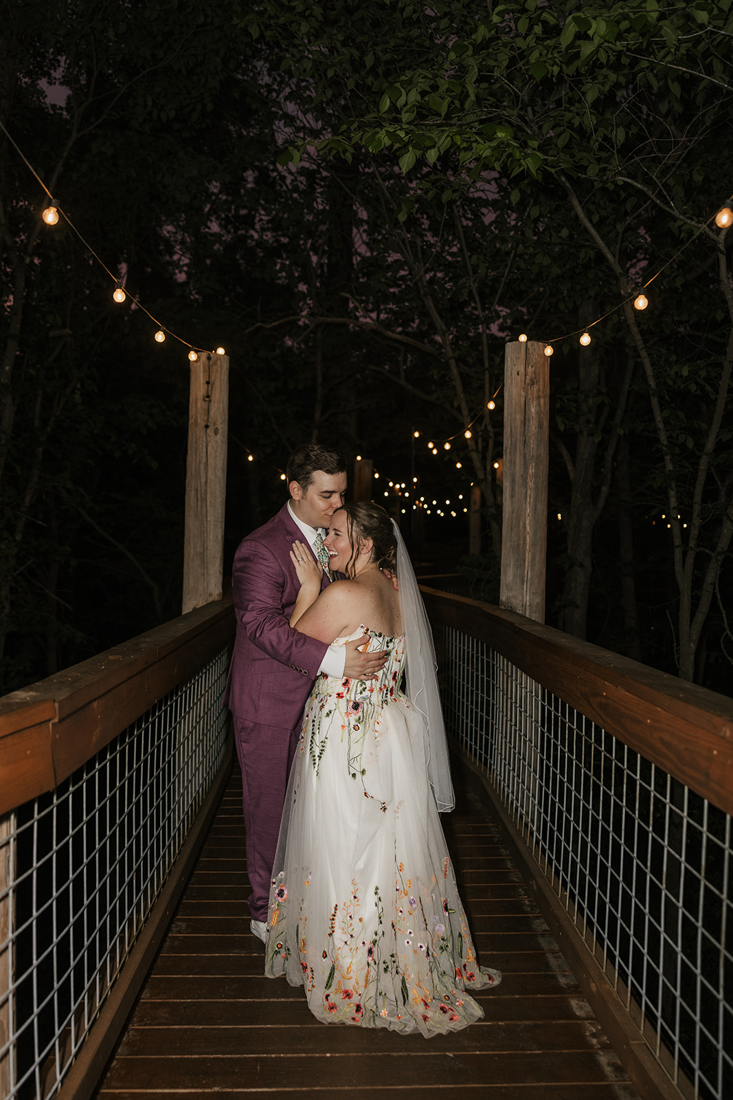 A couple stands on a wooden bridge under string lights at dusk; the person on the left wears a purple suit, and the person on the right wears a white floral dress for their wedding at Merrick Hollow 