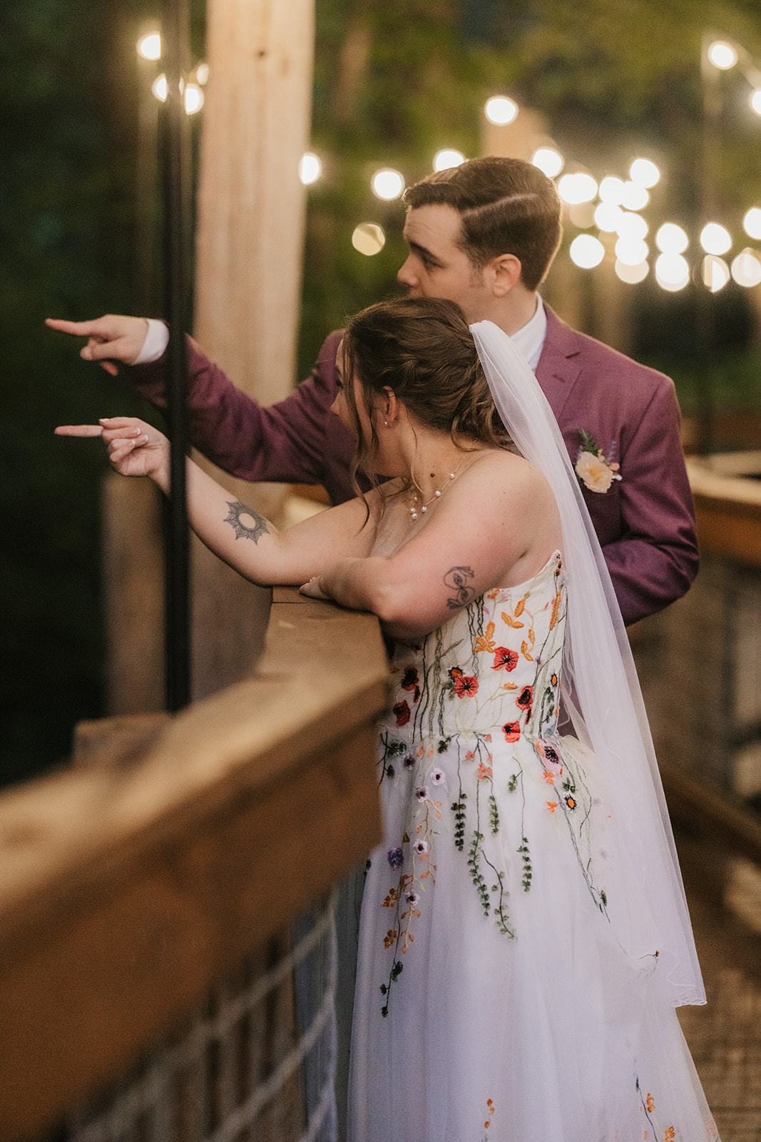 A couple stands on a wooden bridge under string lights at dusk; the person on the left wears a purple suit, and the person on the right wears a white floral dress for their wedding at Merrick Hollow 
