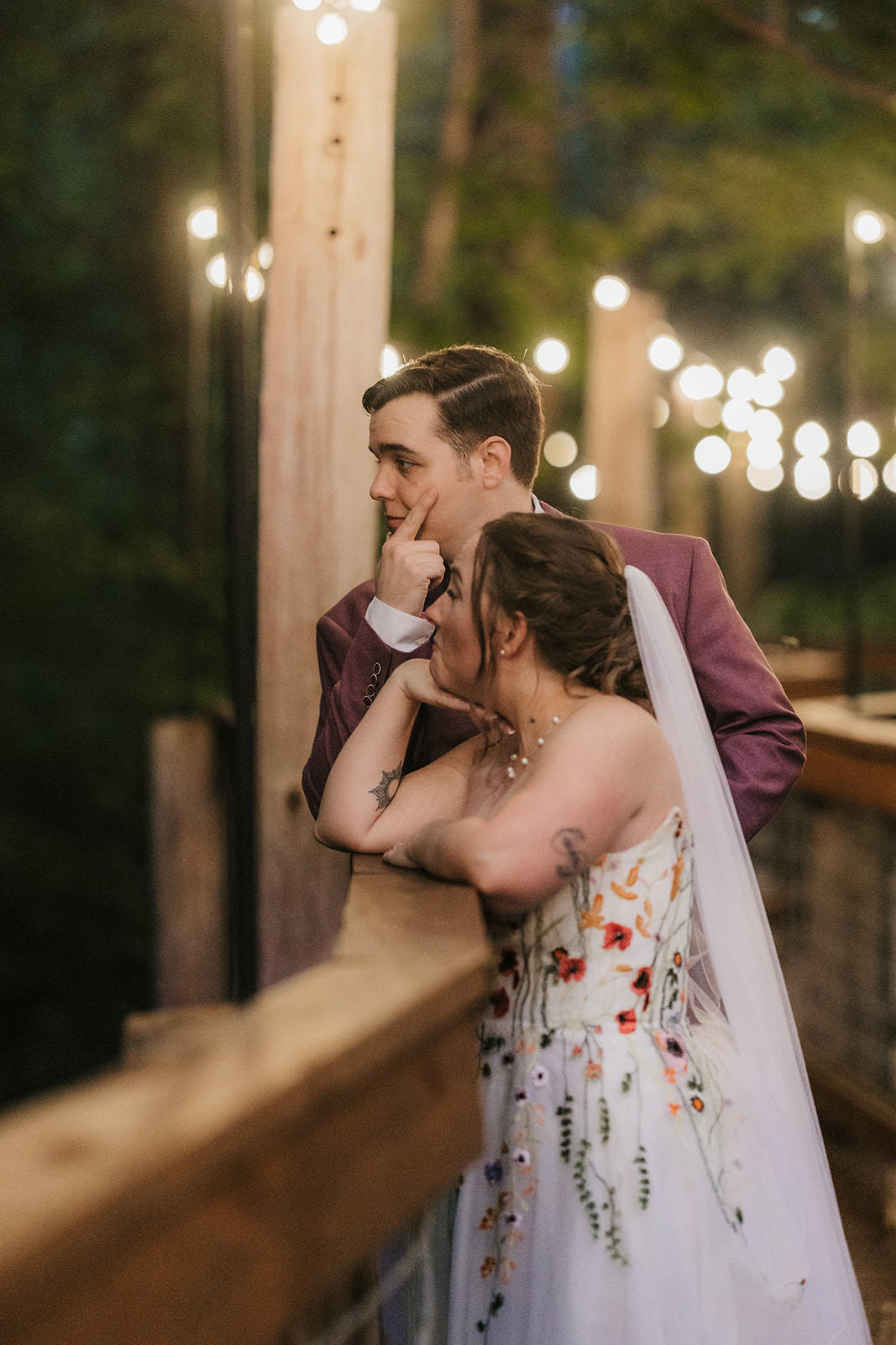 A couple stands on a wooden bridge under string lights at dusk; the person on the left wears a purple suit, and the person on the right wears a white floral dress for their wedding at Merrick Hollow 