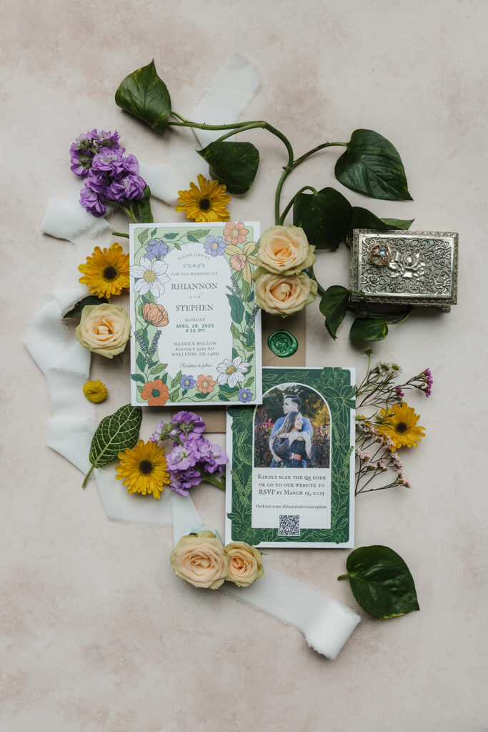 Two floral wedding invitations surrounded by yellow and purple flowers, green leaves, a wax seal, and a decorative box arranged on a light pink surface.