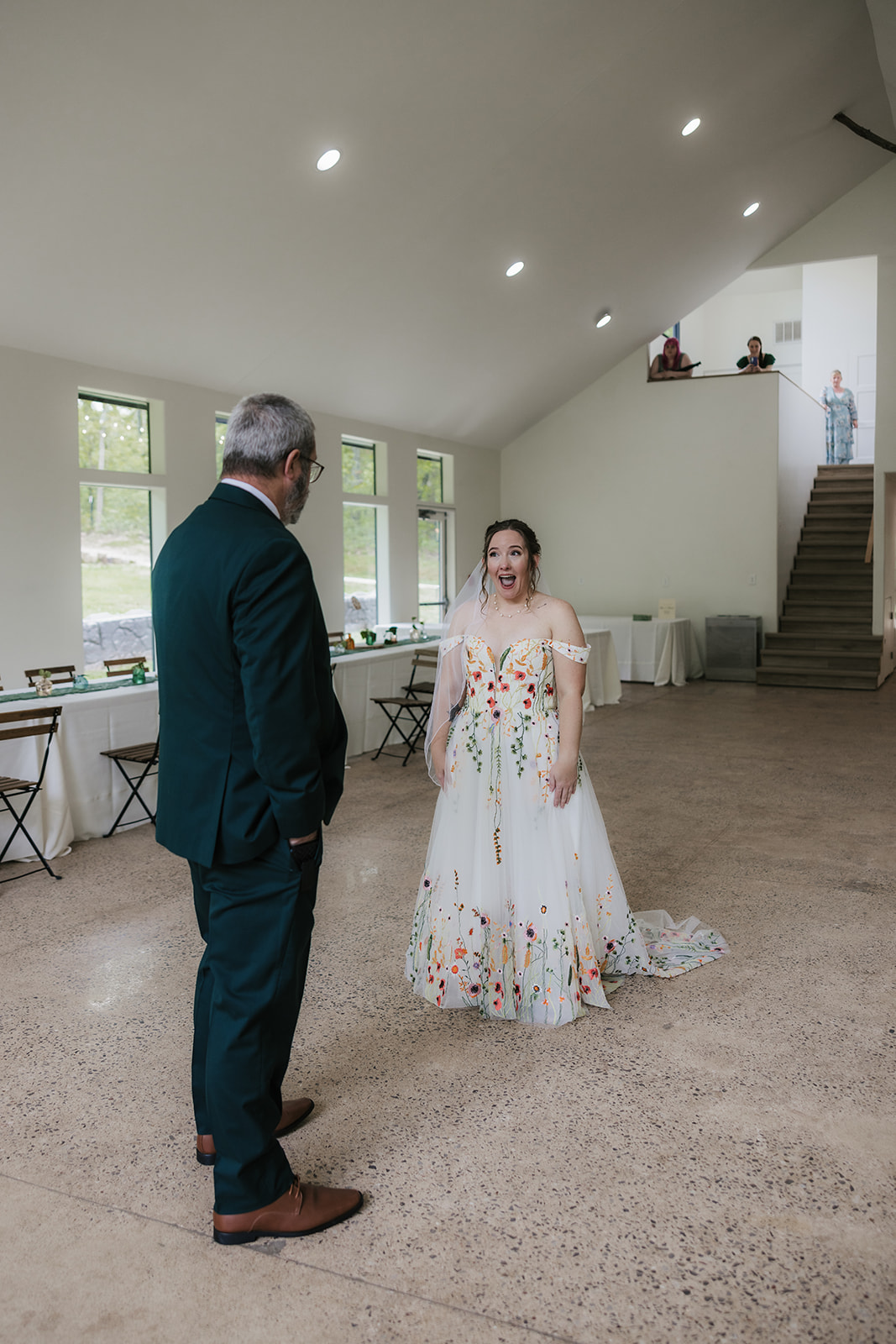 A man in a dark suit stands facing a woman in a white dress with colorful floral patterns in a spacious, well-lit room with tables and chairs.