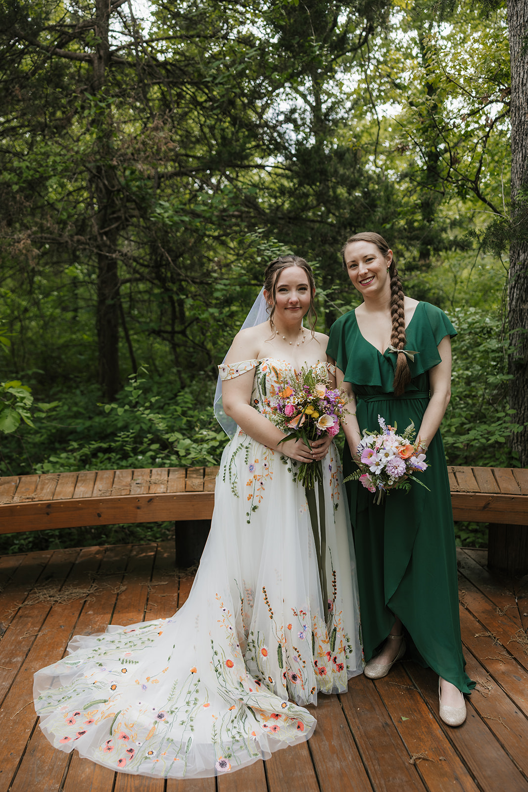A wedding party poses on a wooden deck in a forested area. The group includes the bride and groom, with bridesmaids and groomsmen dressed in formal attire for their wedding at Merrick Hollow