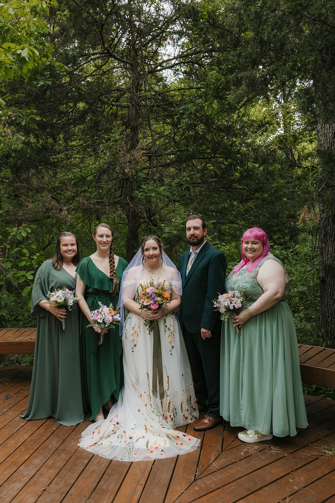 A wedding party poses on a wooden deck in a forested area. The group includes the bride and groom, with bridesmaids and groomsmen dressed in formal attire for their wedding at Merrick Hollow 