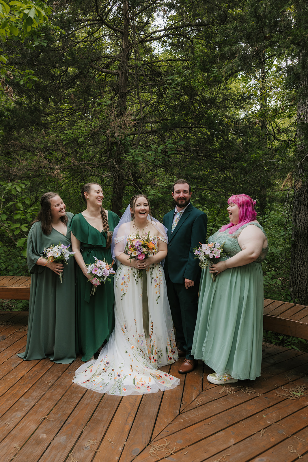 A wedding party poses on a wooden deck in a forested area. The group includes the bride and groom, with bridesmaids and groomsmen dressed in formal attire for their wedding at Merrick Hollow 