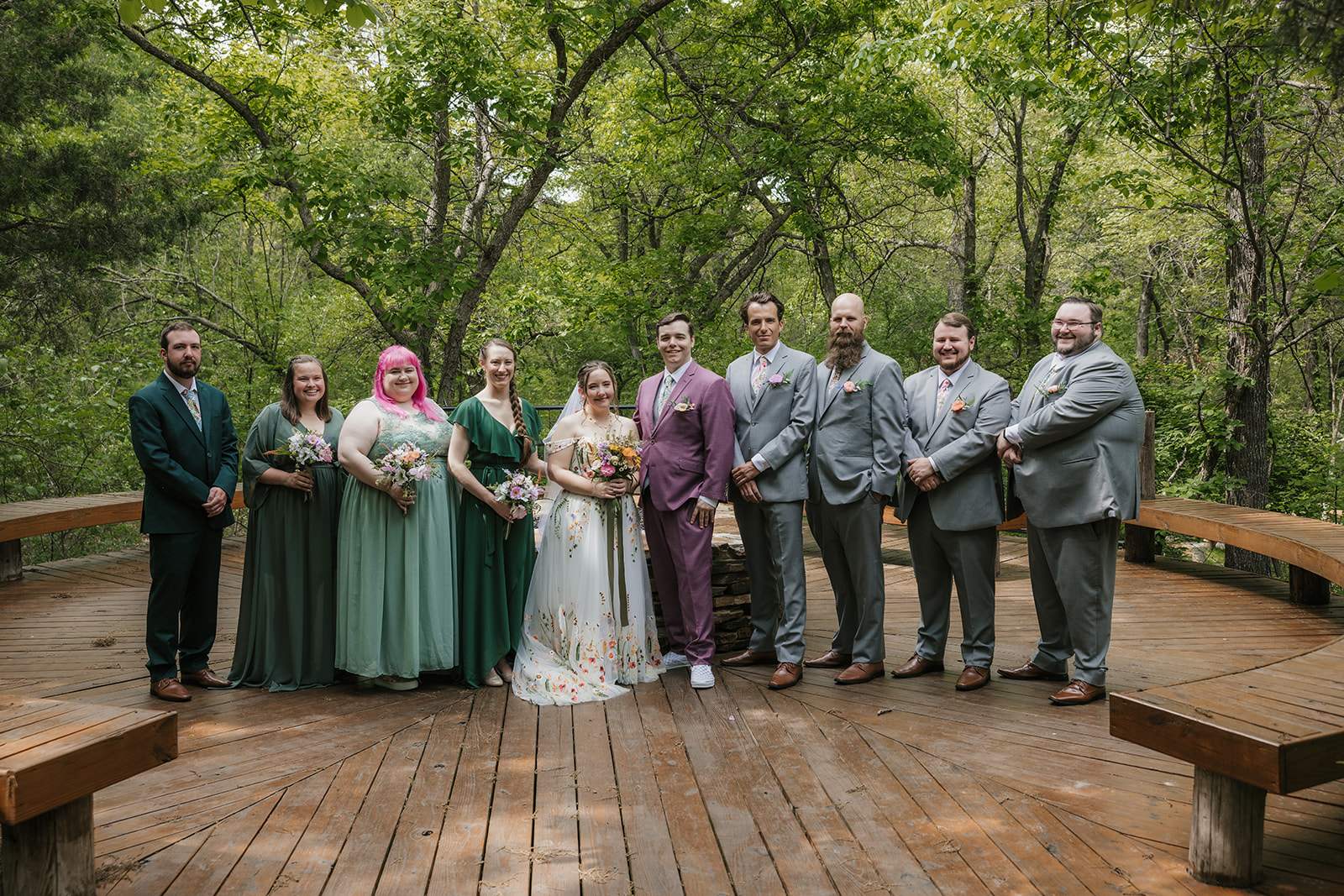 A wedding party poses on a wooden deck in a forested area. The group includes the bride and groom, with bridesmaids and groomsmen dressed in formal attire for their wedding at Merrick Hollow 