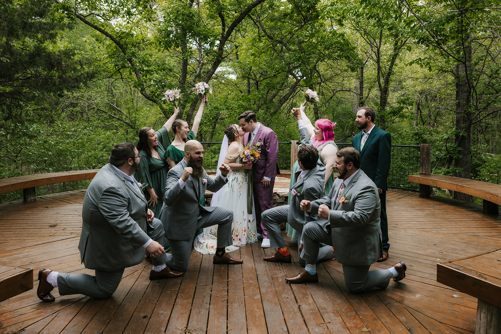 A wedding party poses on a wooden deck in a forested area. The group includes the bride and groom, with bridesmaids and groomsmen dressed in formal attire for their wedding at Merrick Hollow 
