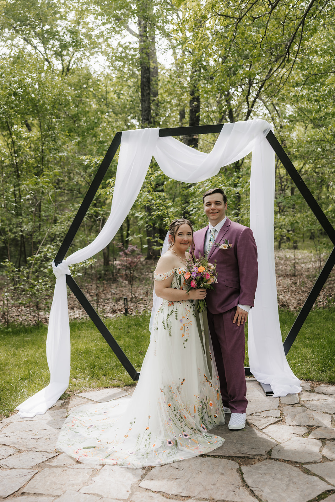 A bride and groom stand together outdoors under a hexagonal arch draped with white fabric, surrounded by greenery.