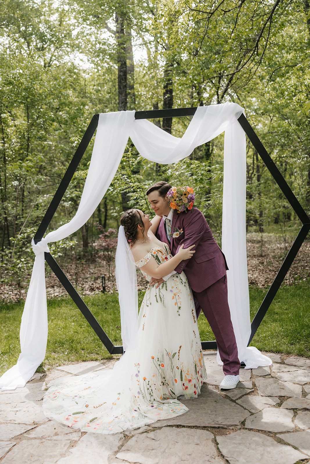 A bride and groom stand together outdoors under a hexagonal arch draped with white fabric, surrounded by greenery.