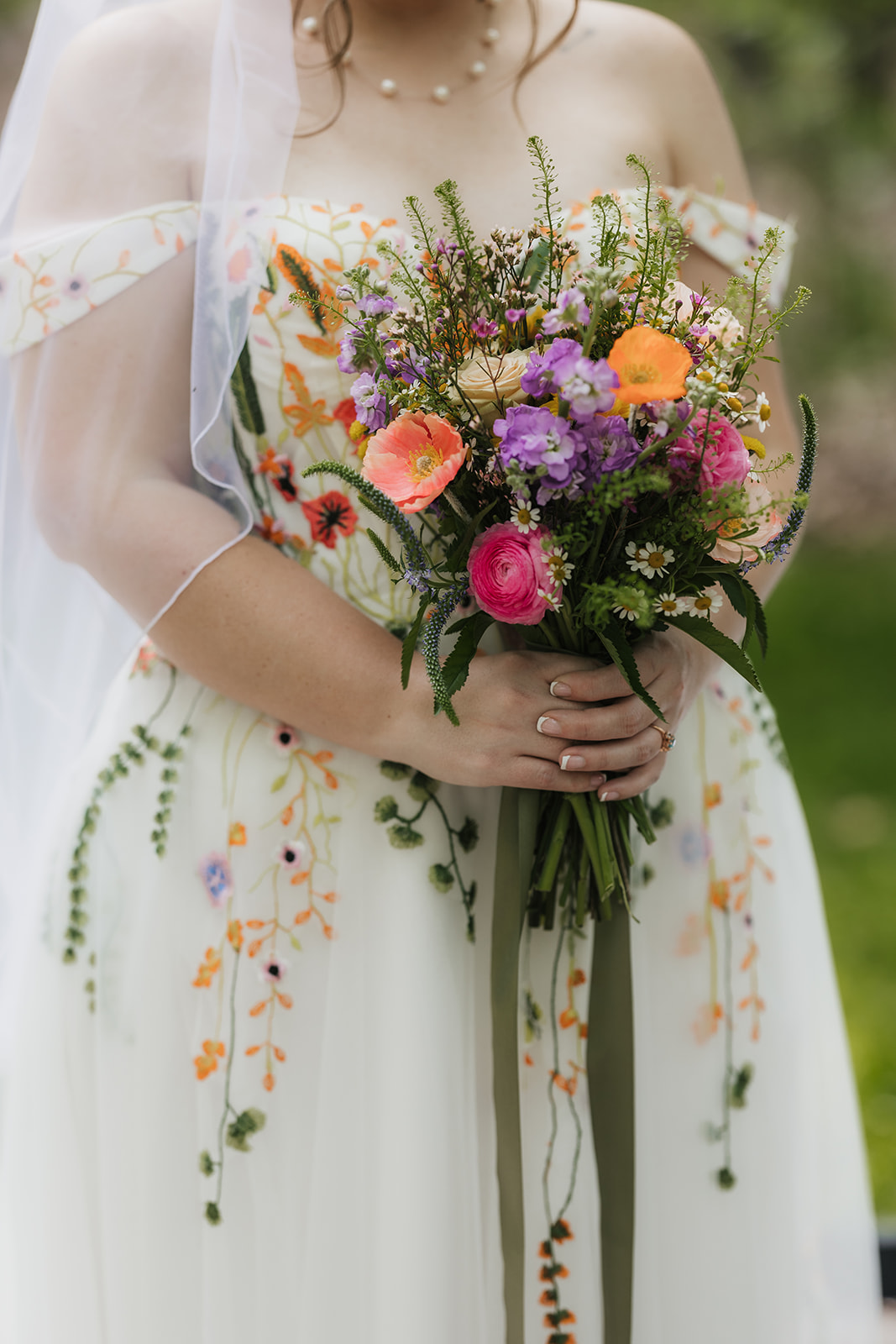 Bride holding a colorful bouquet of flowers, wearing a white dress with floral embroidery, and a sheer veil draped over her shoulders.