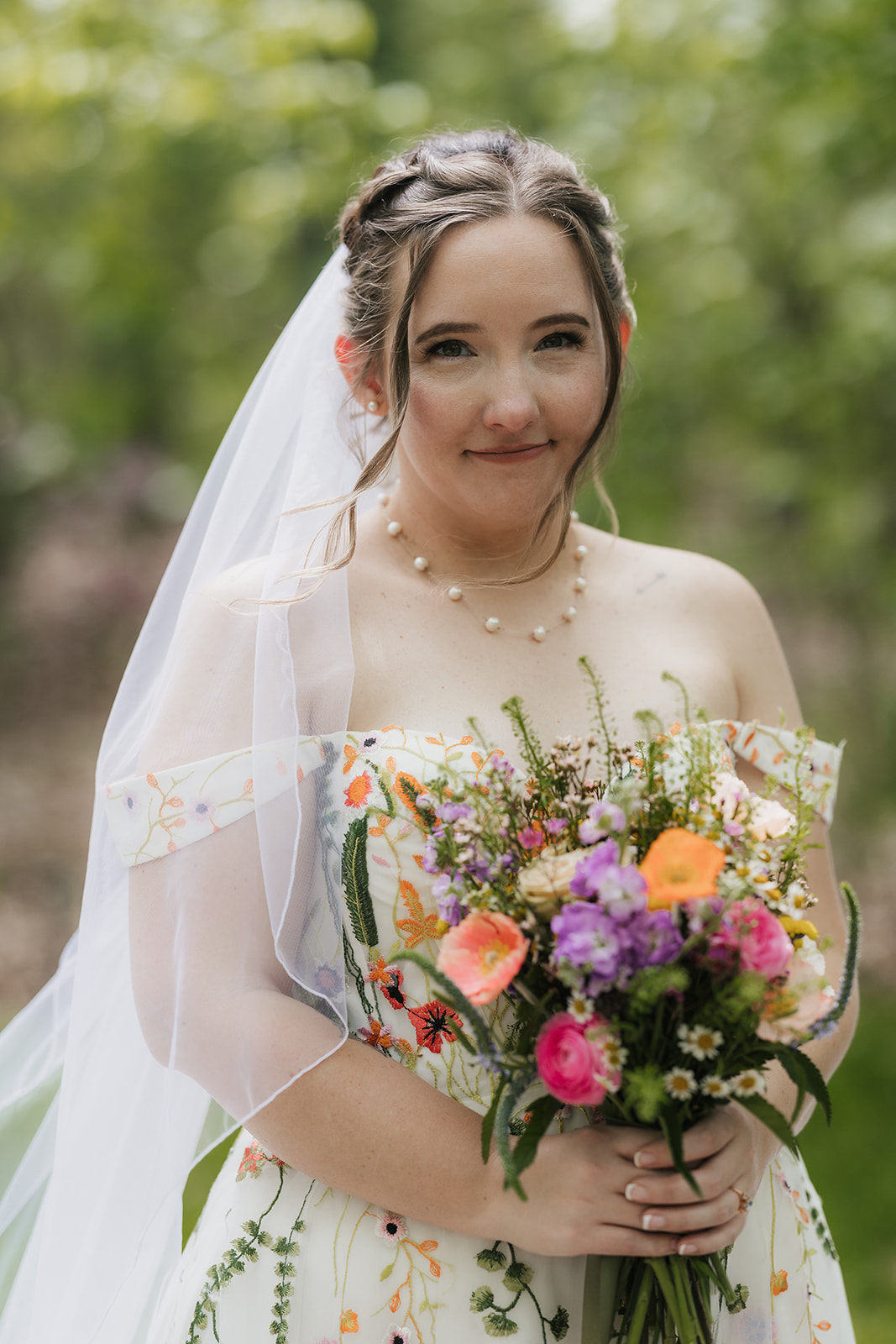 A bride in a white dress with floral embroidery holds a colorful bouquet and stands outdoors, looking at the camera.
