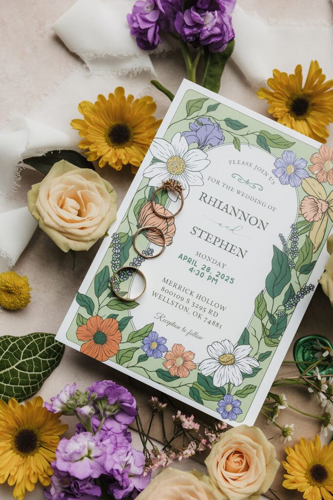 Two floral wedding invitations surrounded by yellow and purple flowers, green leaves, a wax seal, and a decorative box arranged on a light pink surface.
