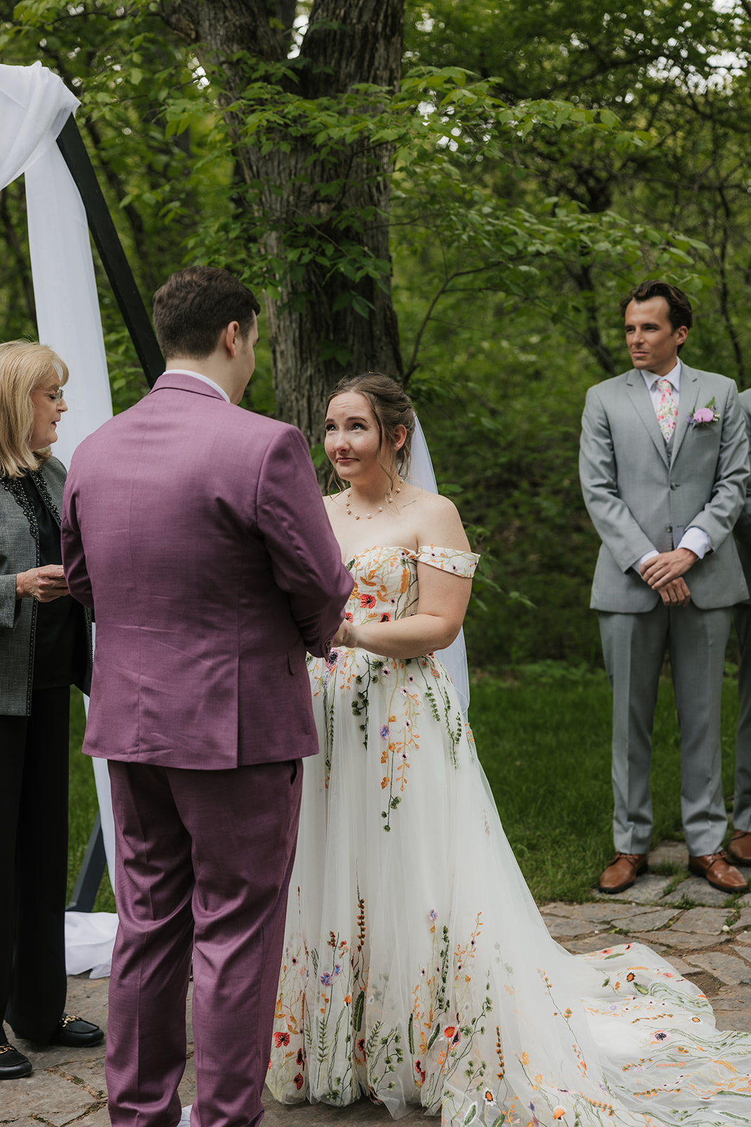 A couple under a geometric arch draped with white fabric during an outdoor wedding ceremony as guests watch and applaud for a wedding at Merrick Hollow Wedding