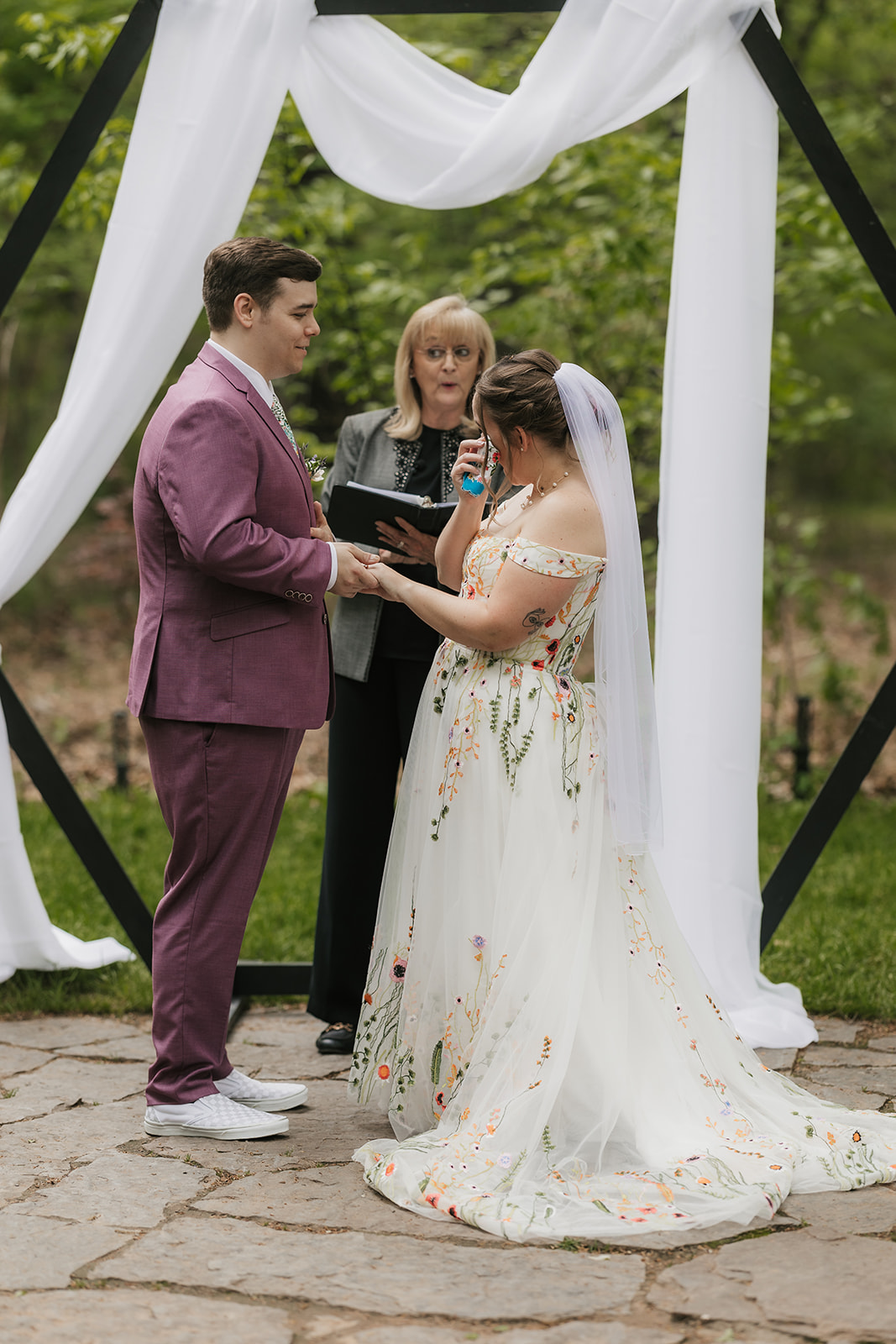 A couple under a geometric arch draped with white fabric during an outdoor wedding ceremony as guests watch and applaud for a wedding at Merrick Hollow Wedding