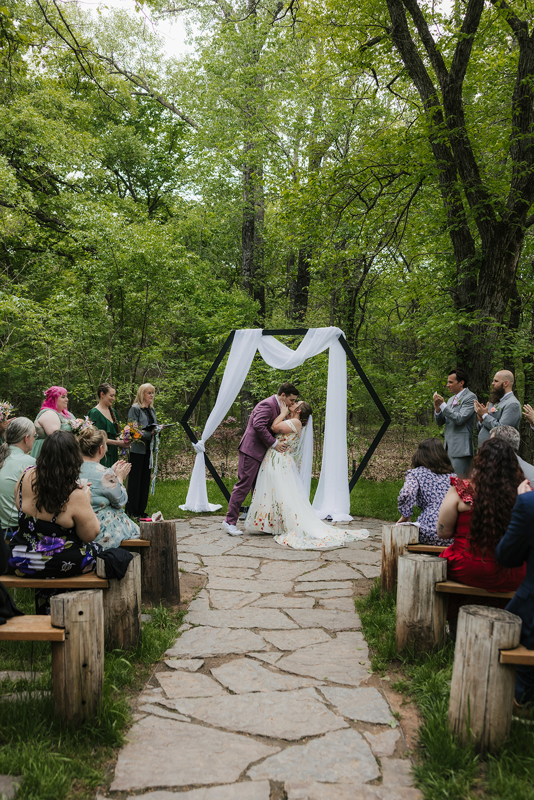 A couple kisses under a geometric arch draped with white fabric during an outdoor wedding ceremony as guests watch and applaud for a wedding at Merrick Hollow Wedding