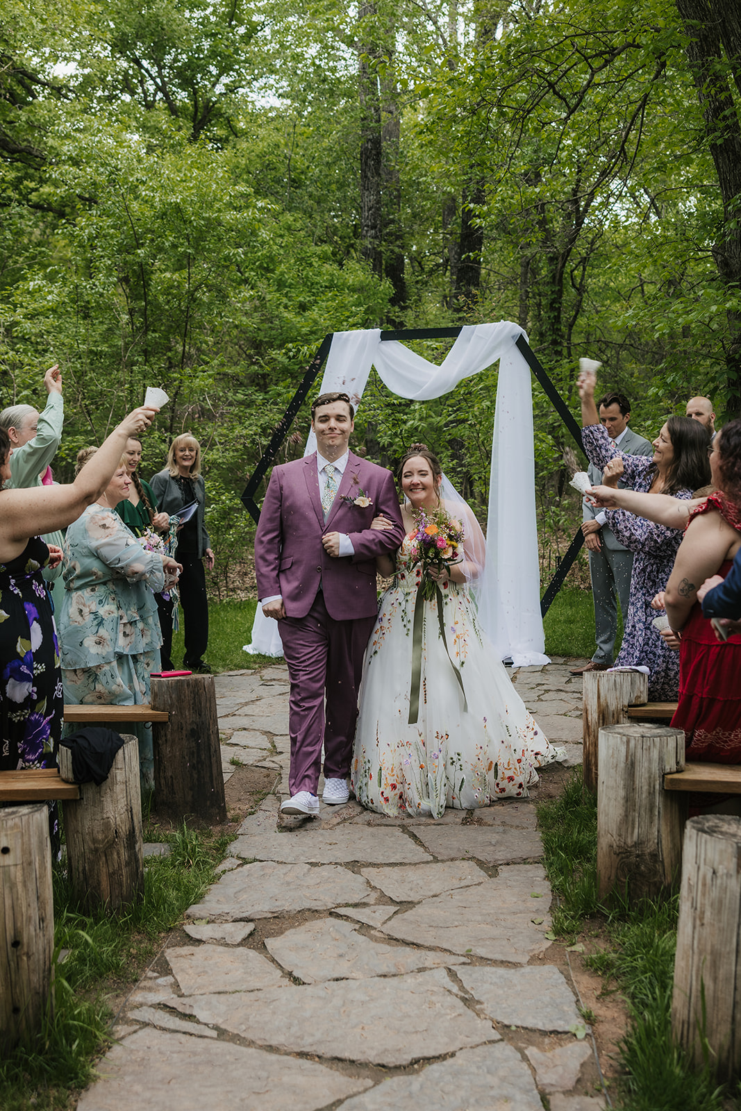 A couple under a geometric arch draped with white fabric during an outdoor wedding ceremony as guests watch and applaud for a wedding at Merrick Hollow Wedding