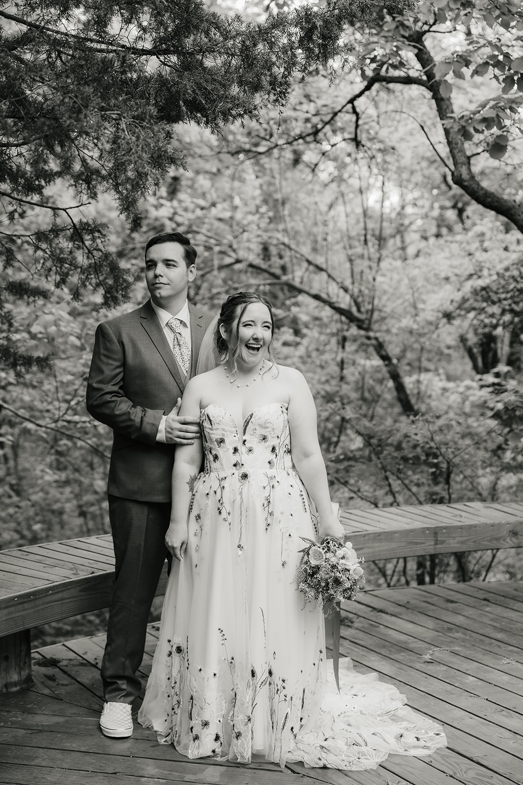 A bride and groom embrace on a wooden deck surrounded by greenery; the groom wears a mauve suit and the bride wears a white dress with floral details and a veil.