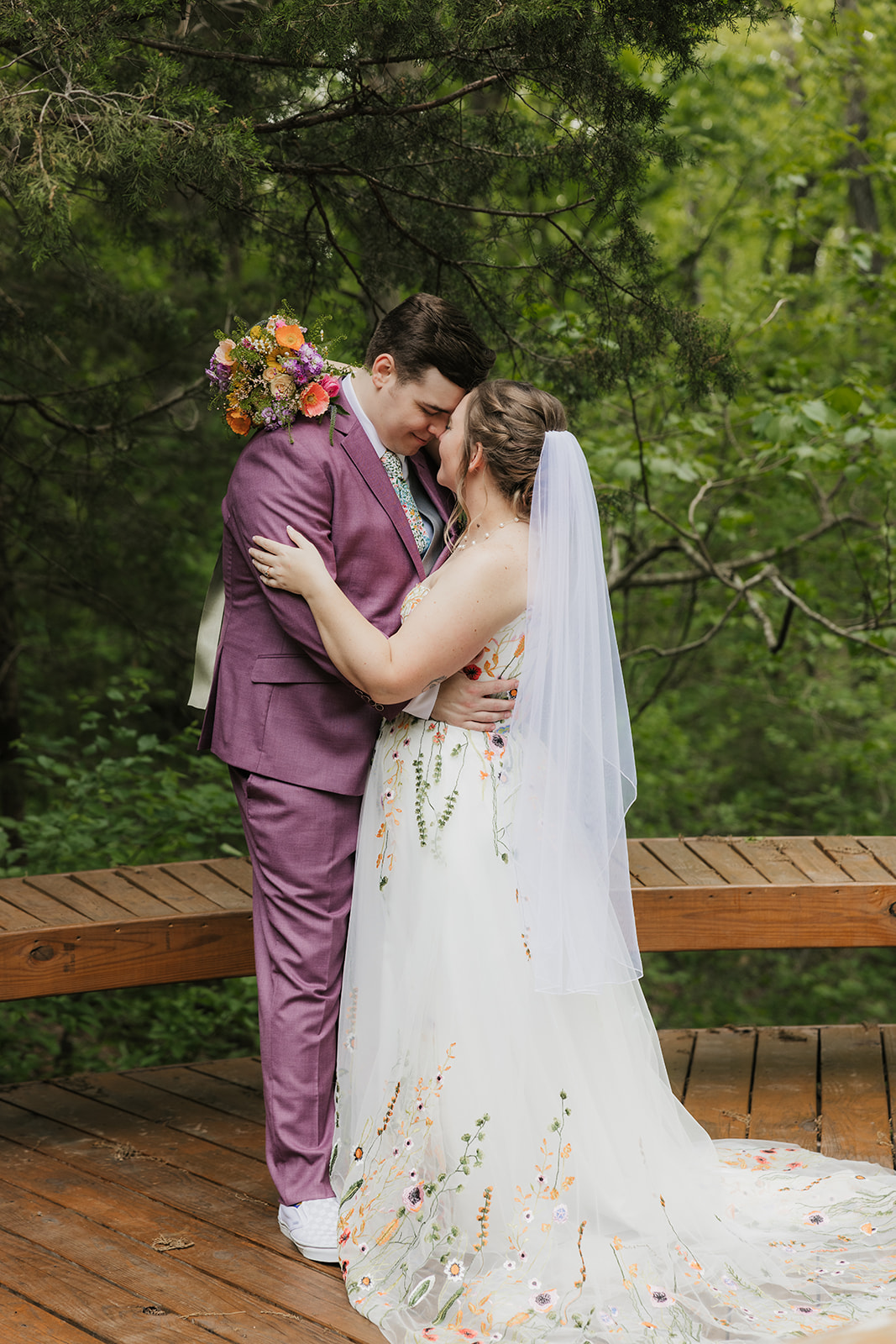 A bride and groom embrace on a wooden deck surrounded by greenery; the groom wears a mauve suit and the bride wears a white dress with floral details and a veil.