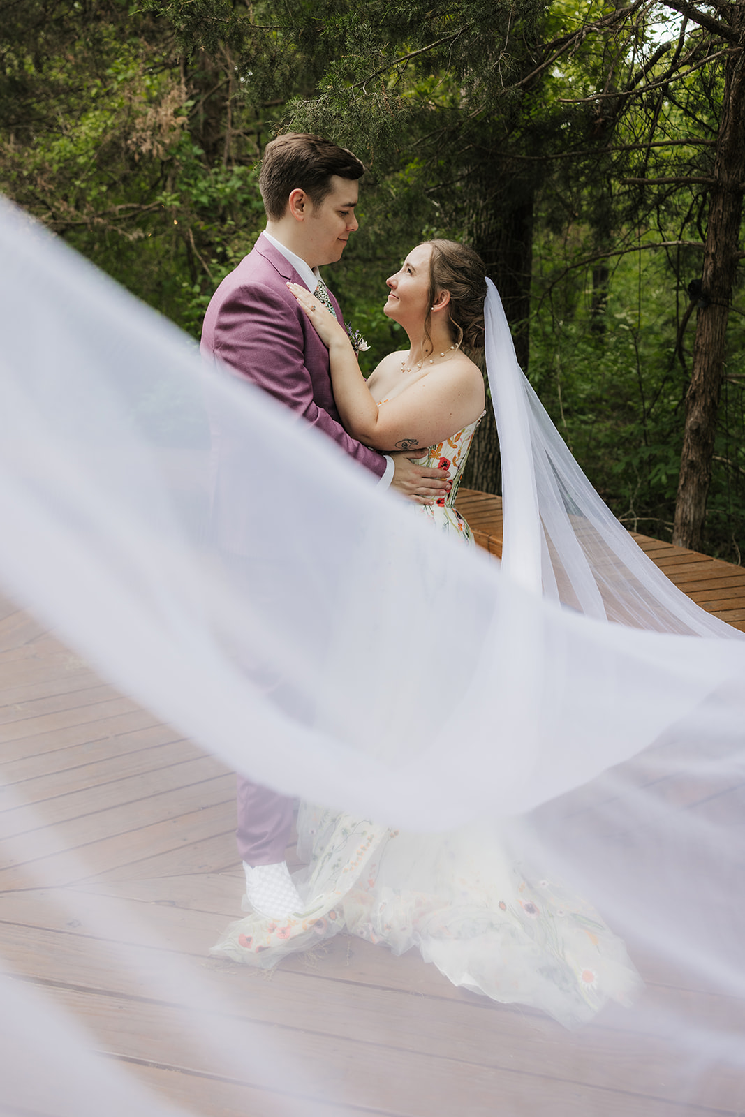 A bride and groom embrace on a wooden deck surrounded by greenery; the groom wears a mauve suit and the bride wears a white dress with floral details and a veil.
