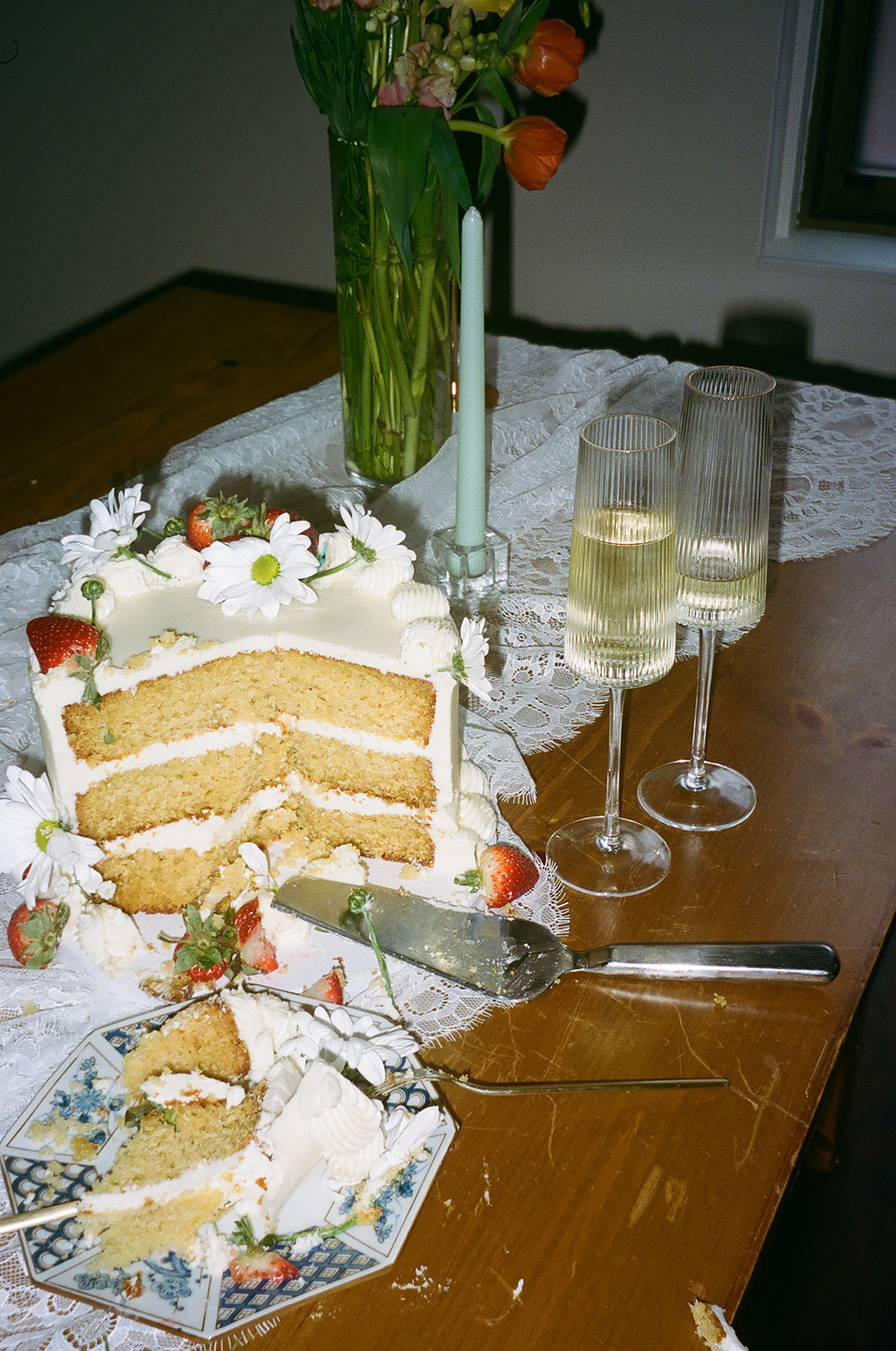 Three-layer vanilla cake with white frosting and strawberries sits on a wooden table with two glasses of champagne, a knife, a candle, and a vase of flowers.