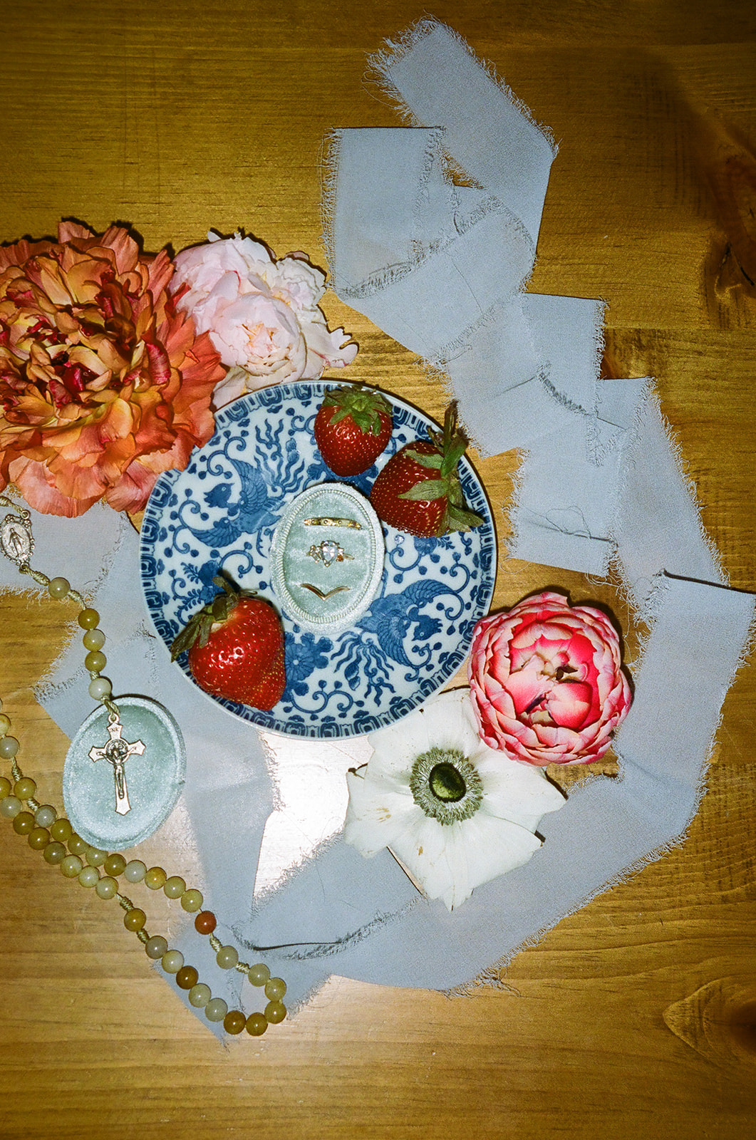 A blue and white decorative plate with strawberries and a porcelain egg, surrounded by flowers, beads, and white fabric on a wooden surface for spring wedding inspo