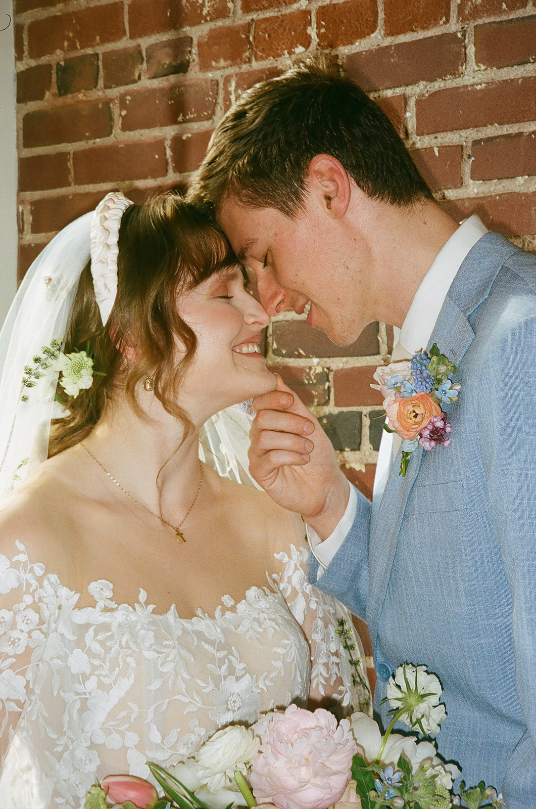 A bride and groom stand close together, smiling and touching foreheads, in front of a brick wall. The bride wears a lace dress and veil; the groom is in a light blue suit with a floral boutonniere for spring wedding inspo