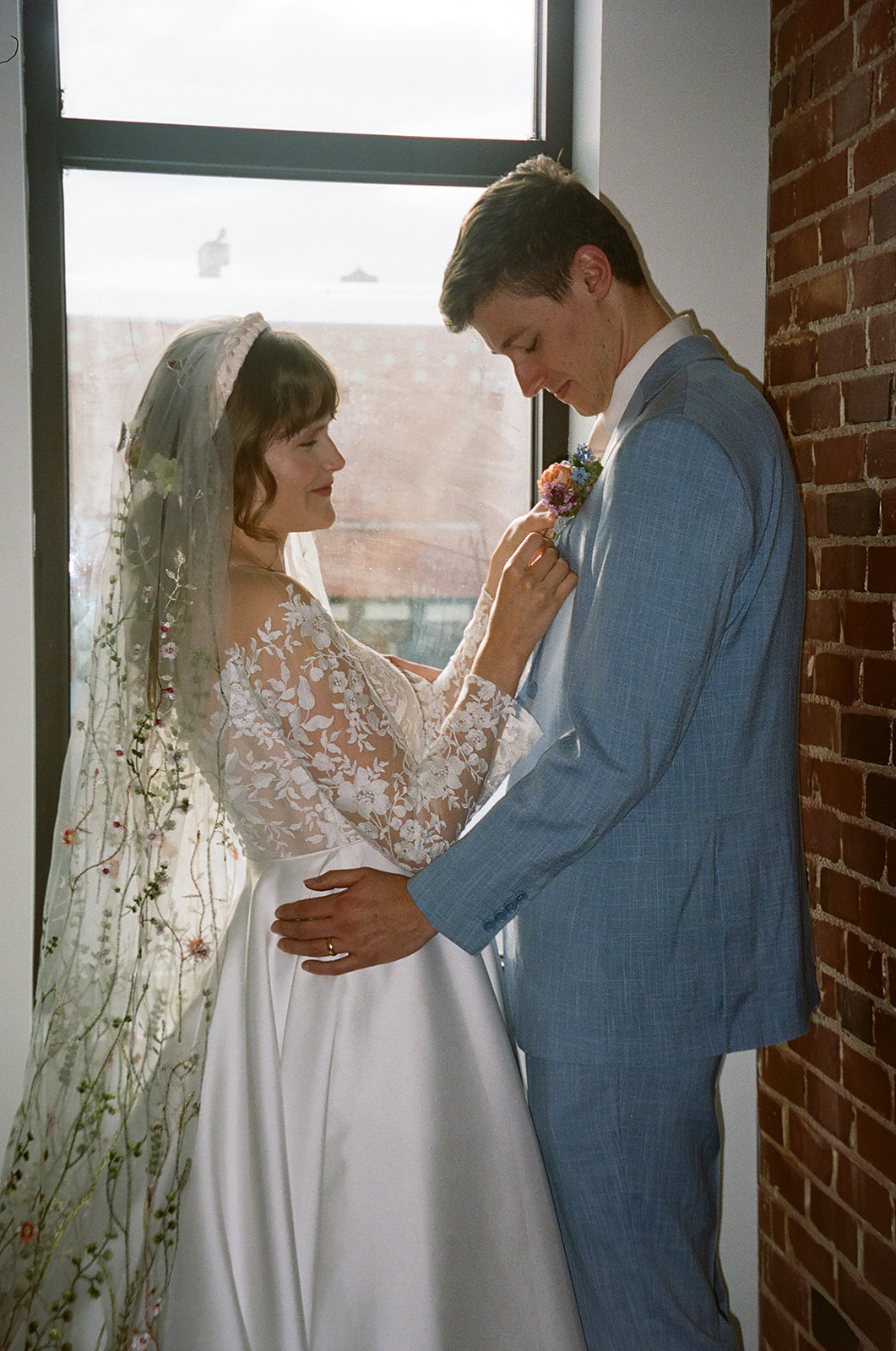 A bride and groom stand close together, smiling and touching foreheads, in front of a brick wall. The bride wears a lace dress and veil; the groom is in a light blue suit with a floral boutonniere for spring wedding inspo