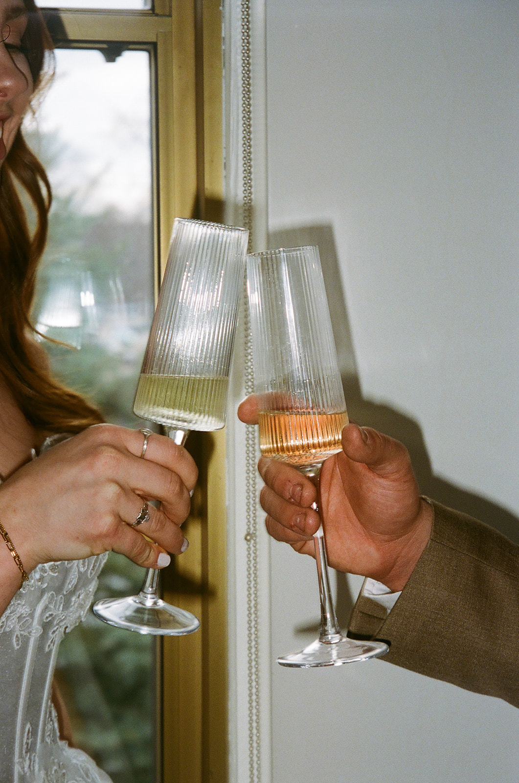 Two people clink champagne glasses near a window, one wearing a white dress and the other in a brown jacket for spring wedding inspo
