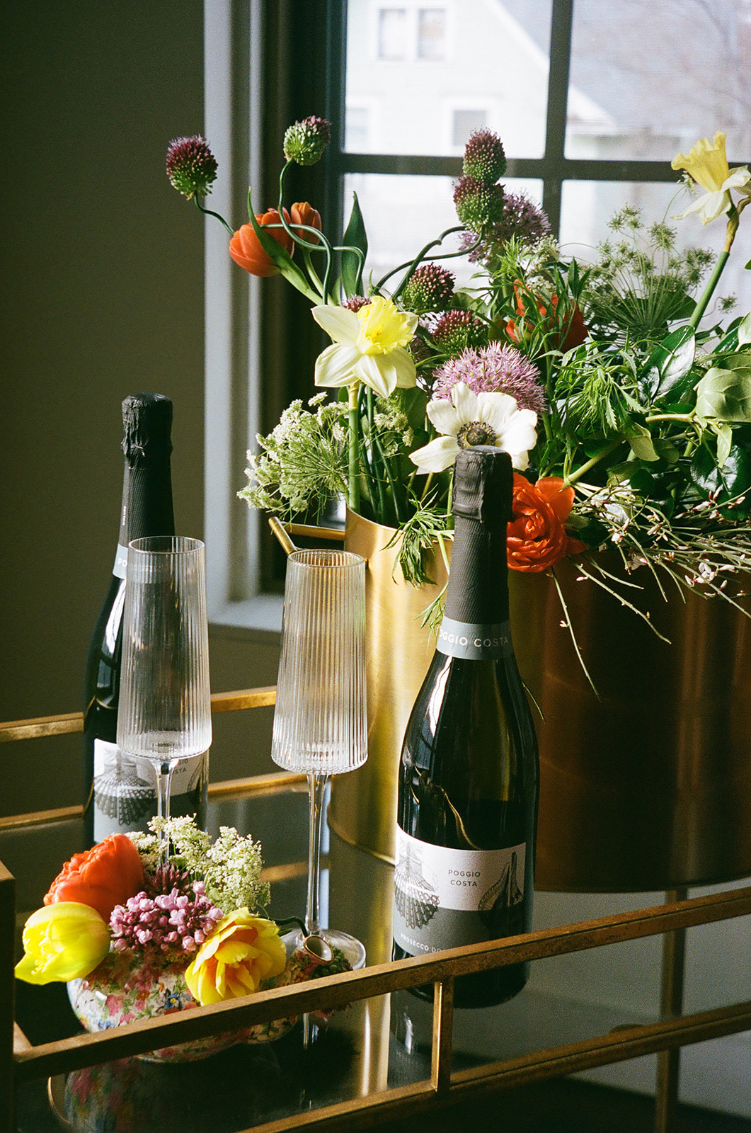 Two people clink champagne glasses near a window, one wearing a white dress and the other in a brown jacket for spring wedding inspo