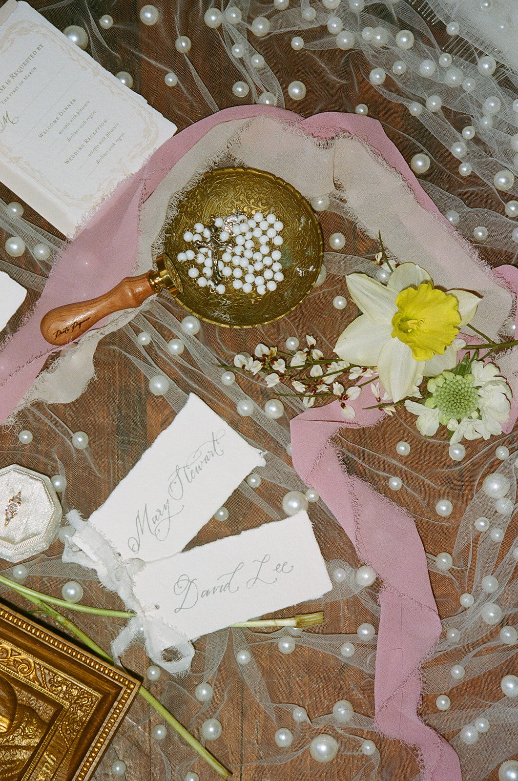 Wedding invitations, stationery, and a religious icon are arranged on a dotted sheer fabric with flowers and ribbons surrounding the items.