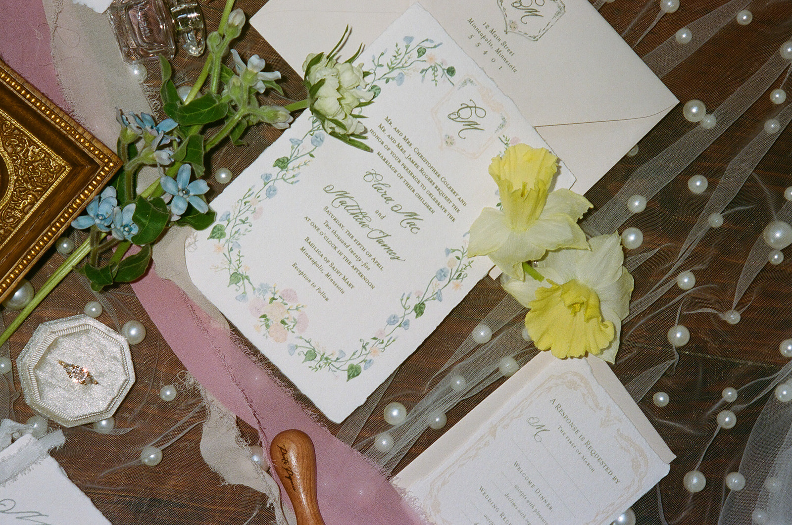 Wedding invitations, stationery, and a religious icon are arranged on a dotted sheer fabric with flowers and ribbons surrounding the items.
