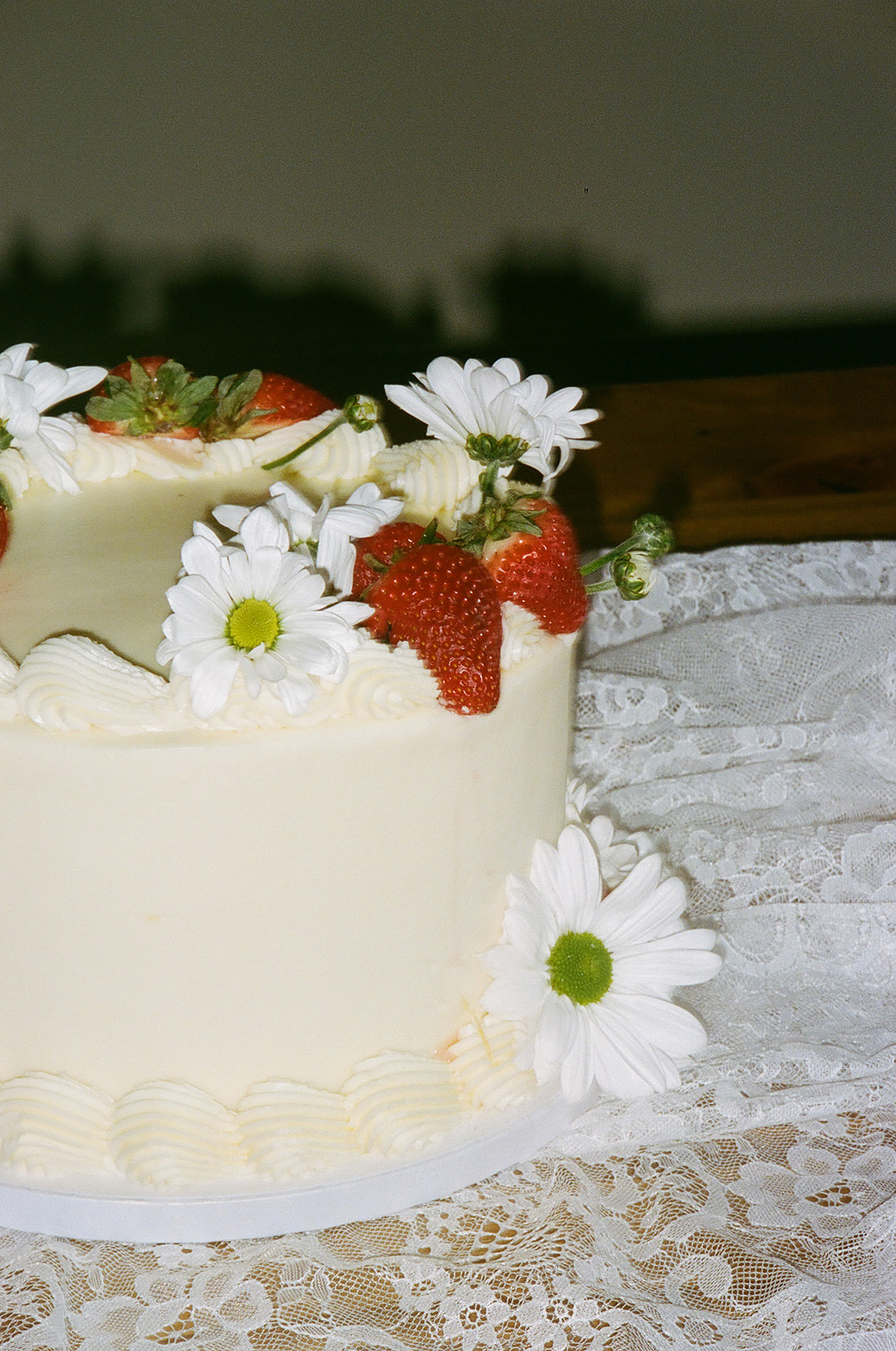 A white frosted cake decorated with fresh strawberries and white daisies sits on a lace tablecloth for spring wedding inspo
