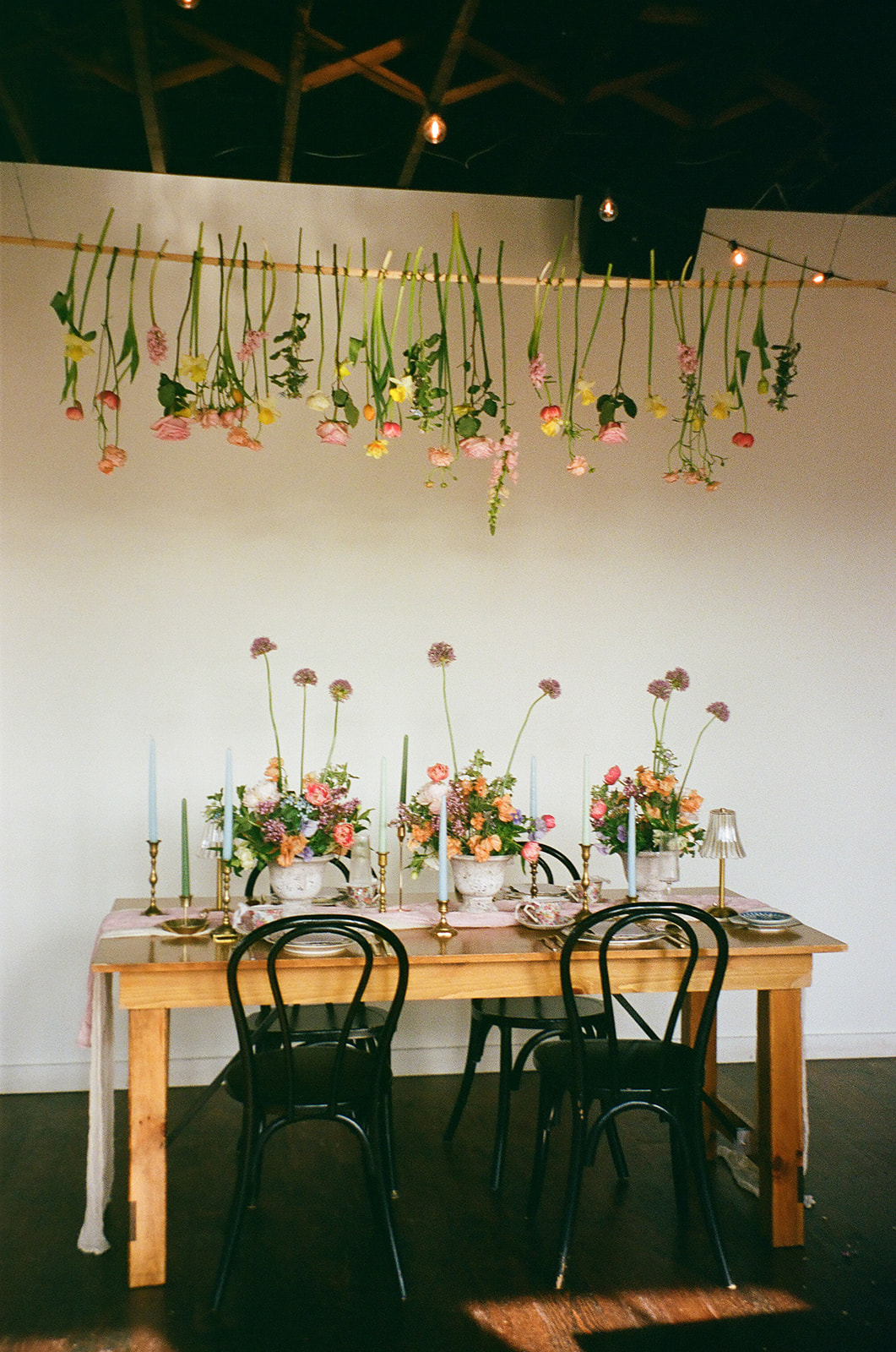 A wooden table set for four with floral centerpieces, black chairs, and flowers hanging upside down above against a white wall.