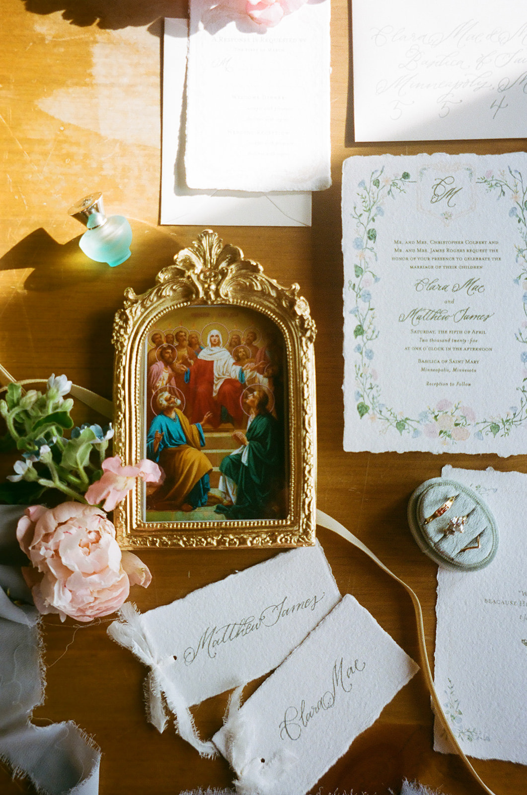 A flat lay of wedding invitations, a framed religious icon, pink and white flowers, a wax seal, and decorative paper on a wooden surface for spring wedding inspo