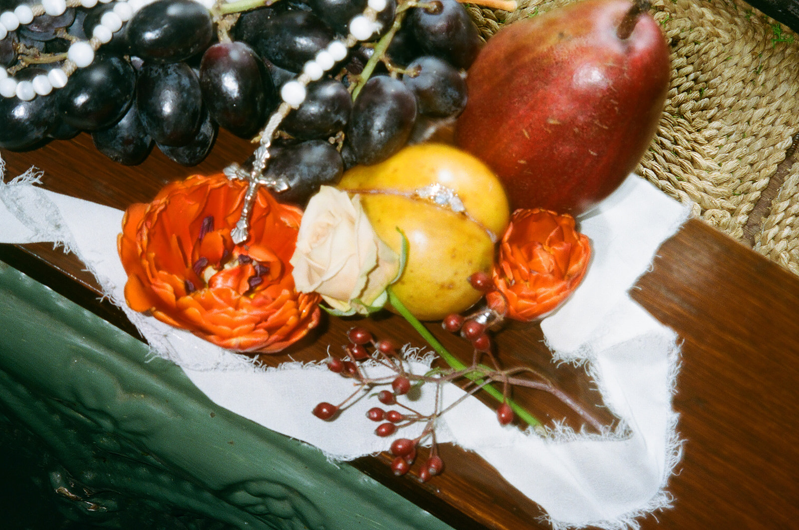 A close-up of assorted fruits, including grapes, a pear, and a yellow fruit, arranged with flowers, berries, and a string of pearls on a white cloth for spring wedding inspo