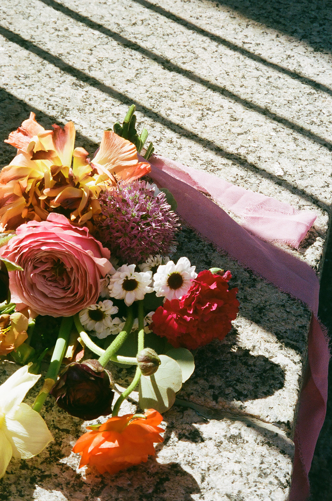 A bouquet of assorted flowers with a pink ribbon lies on a sunlit, textured stone surface with shadows from railings.