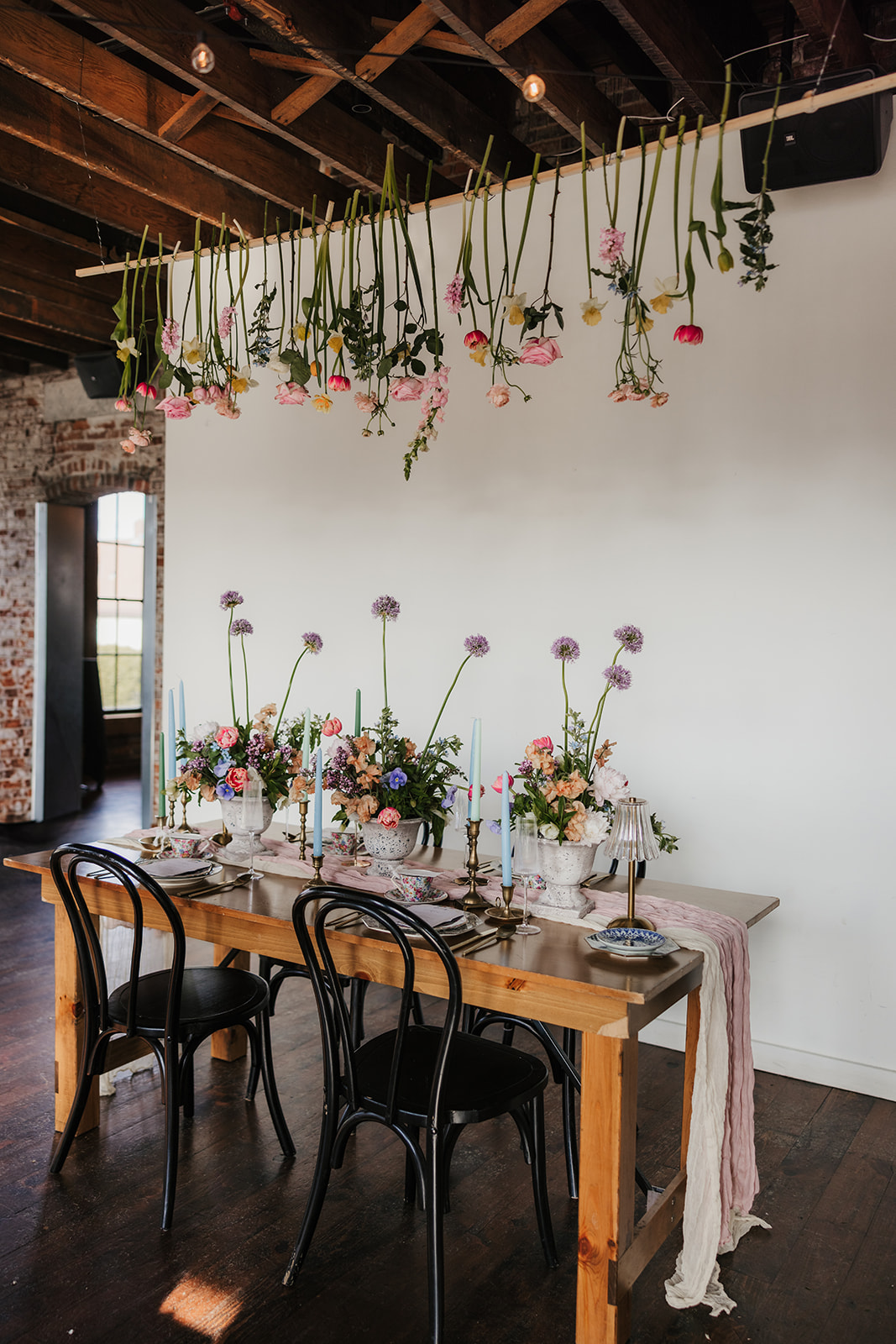 A wooden dining table is set with floral arrangements and candles, with flowers hanging upside down from the ceiling above. Four black chairs surround the table for spring wedding inspo