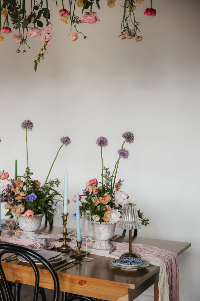A wooden dining table is set with floral arrangements and candles, with flowers hanging upside down from the ceiling above. Four black chairs surround the table for spring wedding inspo