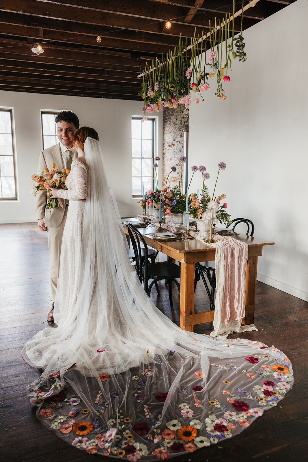 A bride and groom stand together indoors near a decorated table; the bride’s long veil features colorful embroidered flowers spread across the floor.