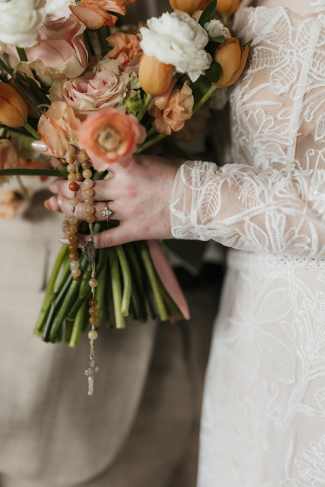 A person in a white lace dress holds a bouquet of orange, white, and pink flowers with a rosary draped around the stems.