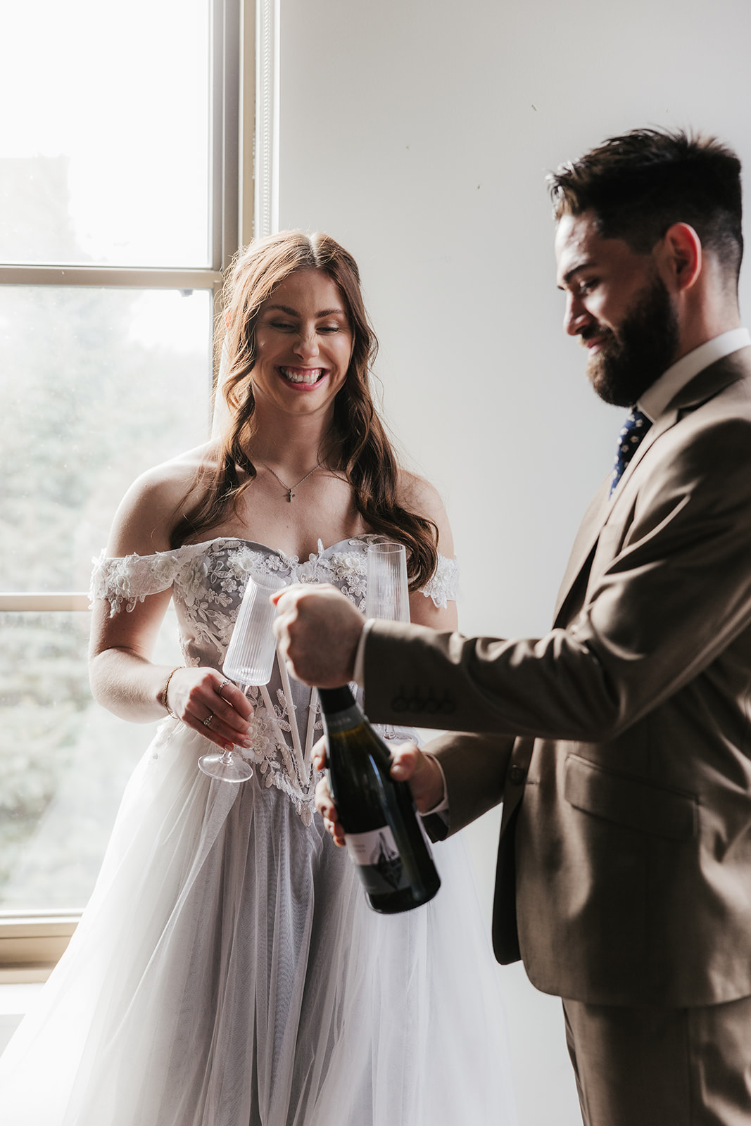 A bride in a white dress and a groom in a tan suit stand by a window, smiling as the groom opens a bottle of champagne and the bride holds a glass.