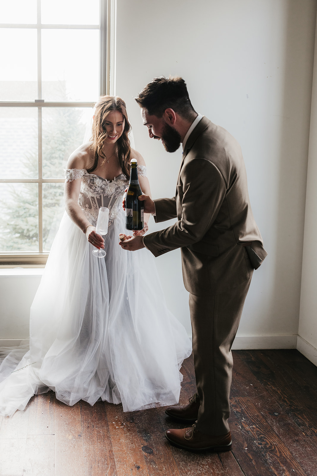 A bride in a white dress and a groom in a tan suit stand by a window, smiling as the groom opens a bottle of champagne and the bride holds a glass.