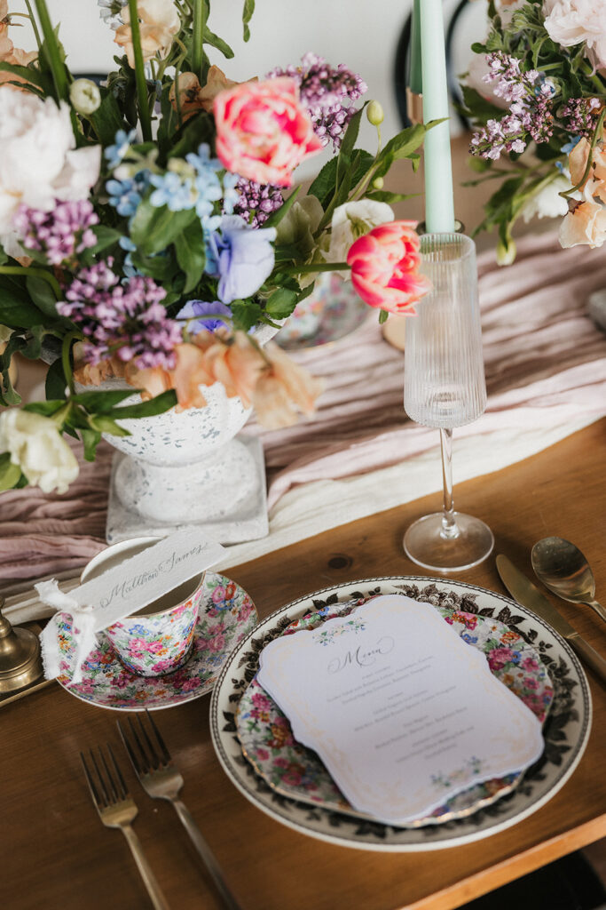A wooden dining table is set with floral arrangements and candles, with flowers hanging upside down from the ceiling above. Four black chairs surround the table for spring wedding inspo