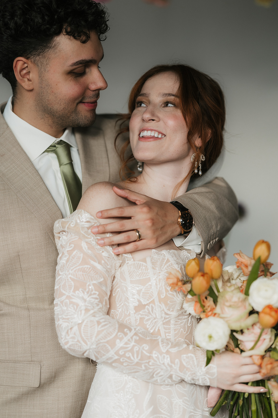 A couple poses together; the man in a tan suit embraces the woman in a lace wedding dress holding a bouquet of flowers.