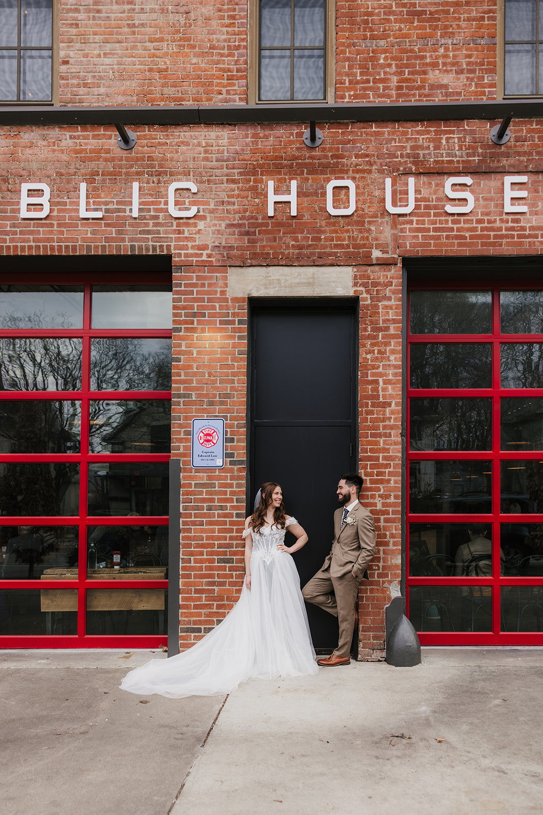 A bride and groom face each other affectionately, with the groom gently holding the bride’s chin, standing in front of a brick wall.