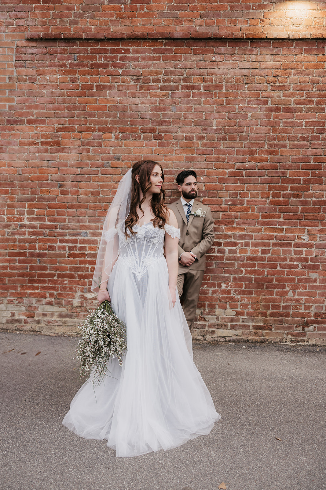 A bride and groom face each other affectionately, with the groom gently holding the bride’s chin, standing in front of a brick wall.