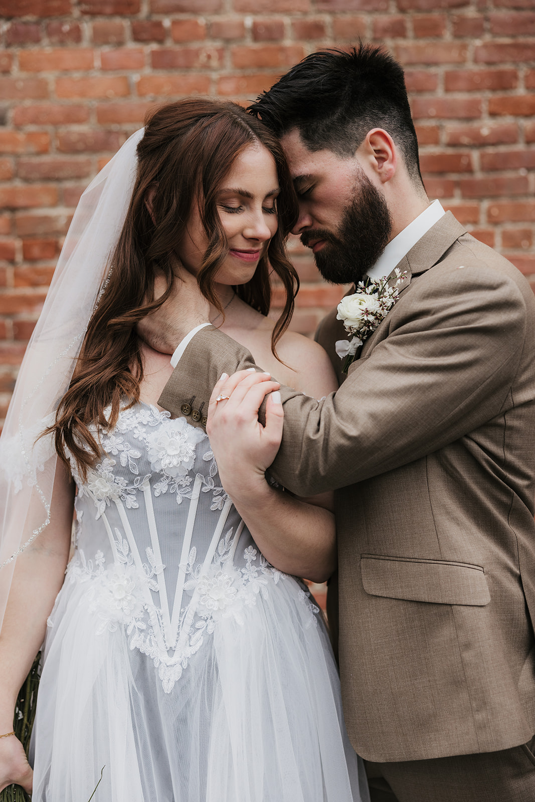 A bride and groom face each other affectionately, with the groom gently holding the bride’s chin, standing in front of a brick wall.
