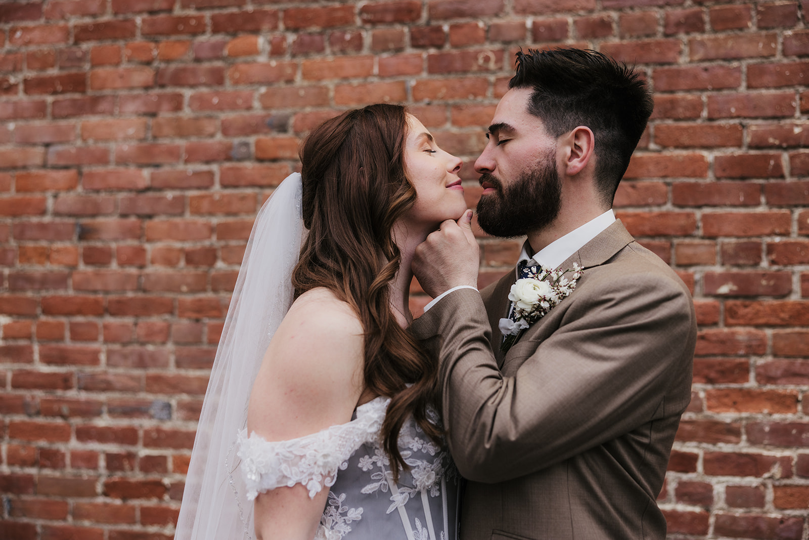 A bride and groom face each other affectionately, with the groom gently holding the bride’s chin, standing in front of a brick wall.